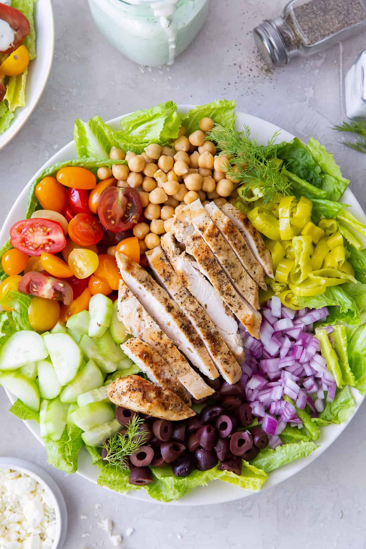 A top down shot of Mediterranean Chicken Salad in a white bowl next to a pepper shaker, feta in a small bowl, and salad dressing.