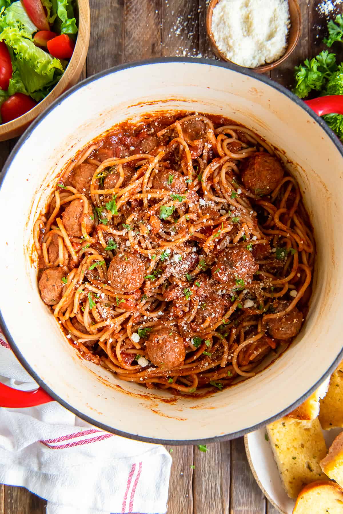 A top down shot of spaghetti and meatballs in a red Dutch oven next to a salad and small bowl of Parmesan.