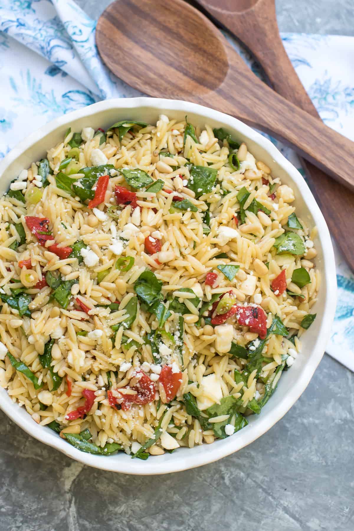 A top down shot of a bowl of orzo pasta salad with spinach and feta next to wooden salad spoons and a blue and white cloth.