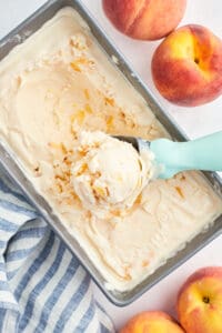 An ice cream scoop with a blue handle scooping frozen peach yogurt from a metal loaf pan.