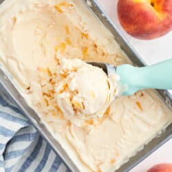 An ice cream scoop with a blue handle scooping frozen peach yogurt from a metal loaf pan.
