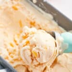 An ice cream scoop with a blue handle scooping frozen peach yogurt from a metal loaf pan.