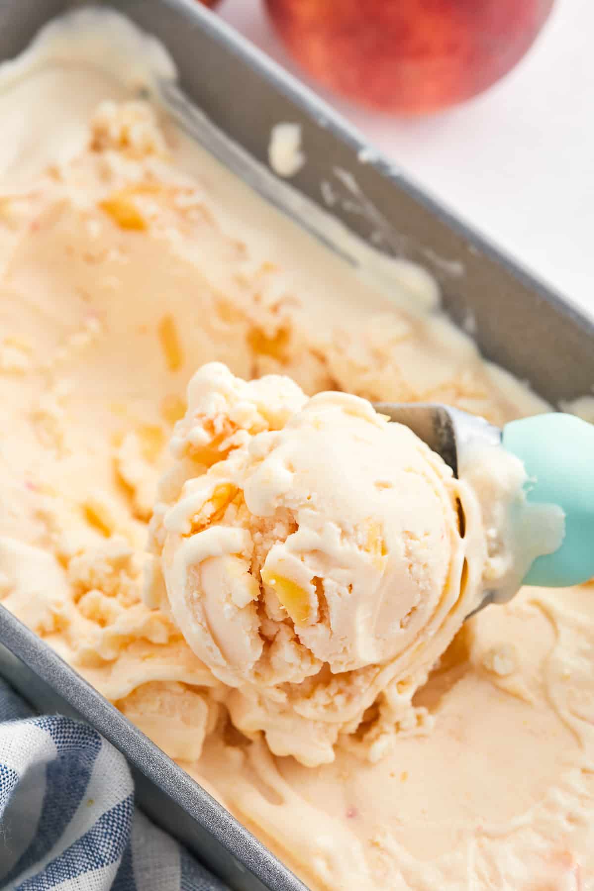 An ice cream scoop with a blue handle scooping frozen peach yogurt from a metal loaf pan.