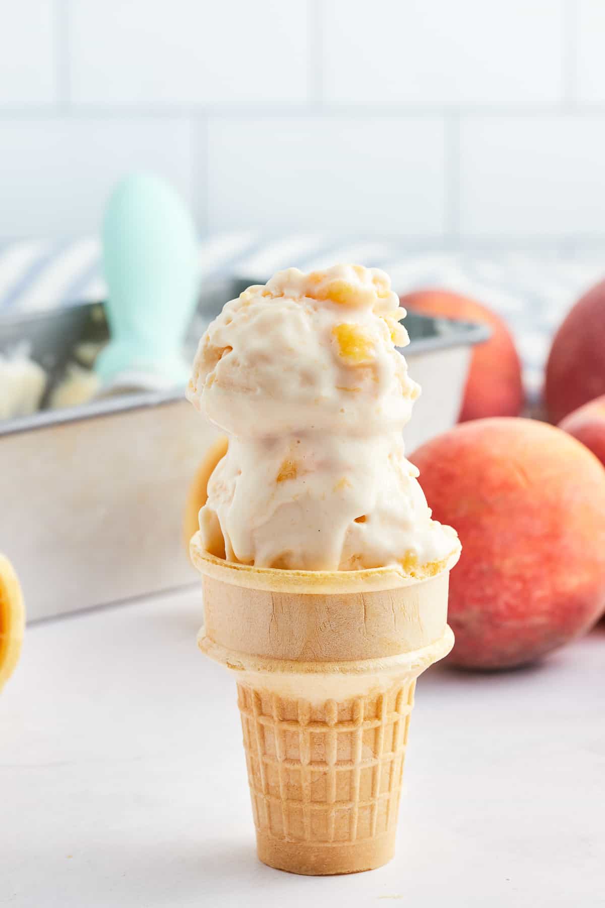 Two scoops of peach frozen yogurt on an ice cream cone in front of fresh peaches.