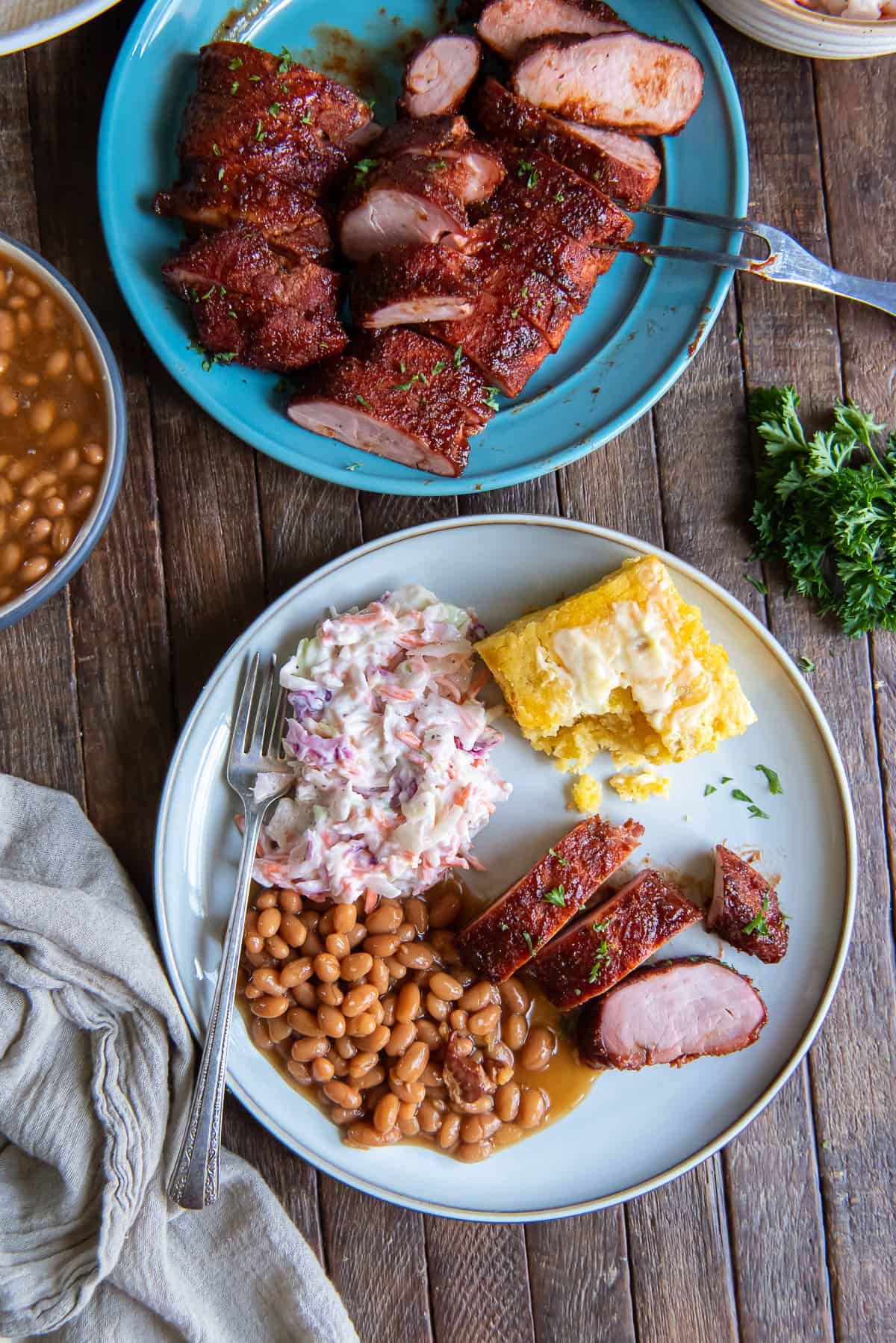 A top down shot of slices of smoked pork tenderloin, cornbread, beans and coleslaw on a white dinner plate.