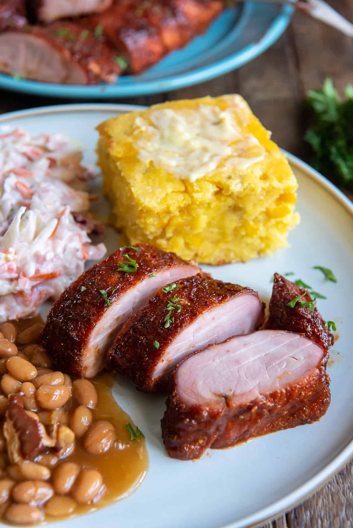 Slices of smoked pork tenderloin, cornbread, beans and coleslaw on a white dinner plate.