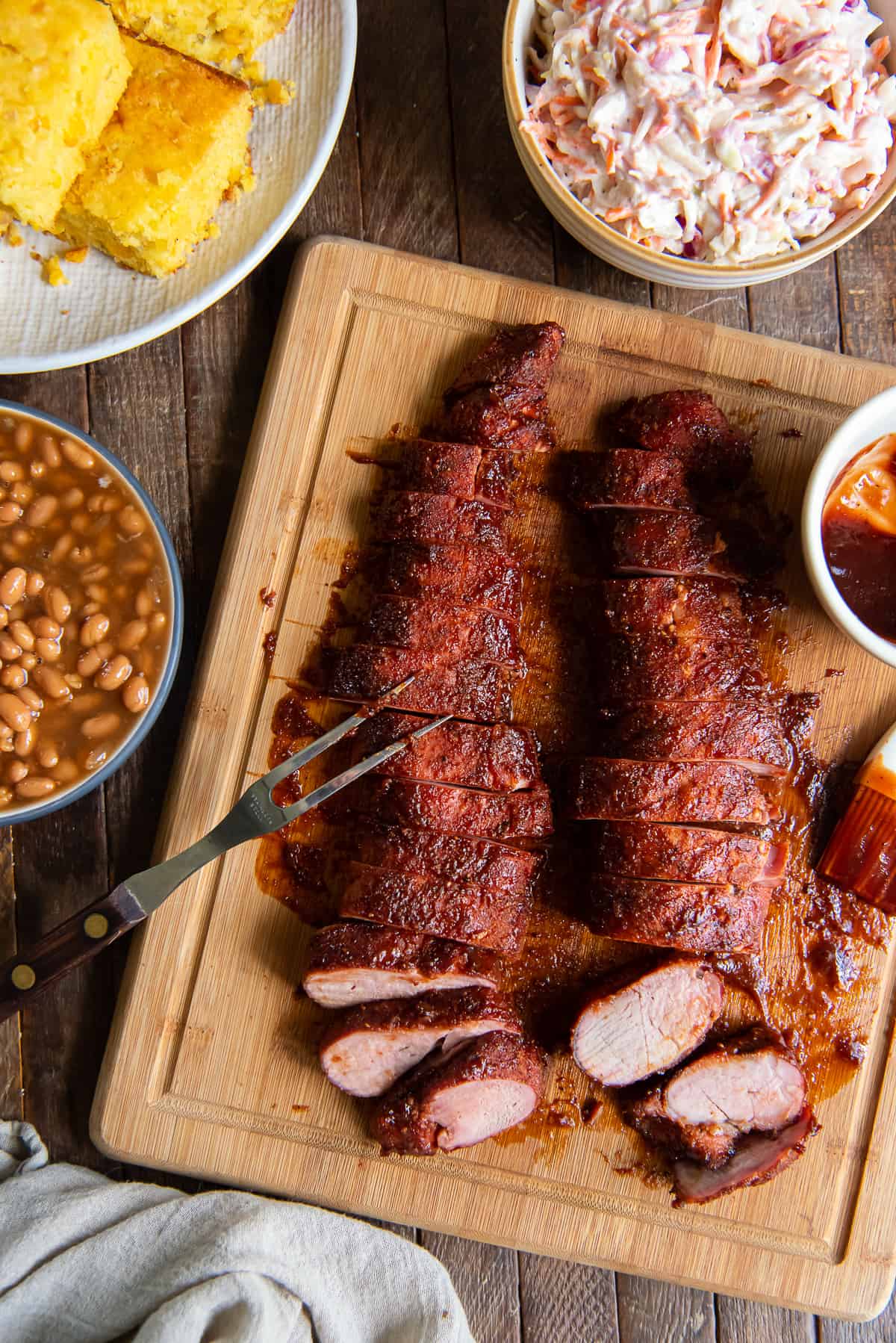 Sliced smoked pork tenderloin on a cutting board next to a platter of cornbread, a bowl of beans, and coleslaw.
