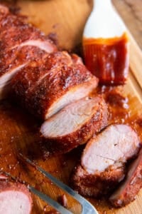 Sliced pork tenderloin on a cutting board with a pastry brush coated in barbecue sauce.