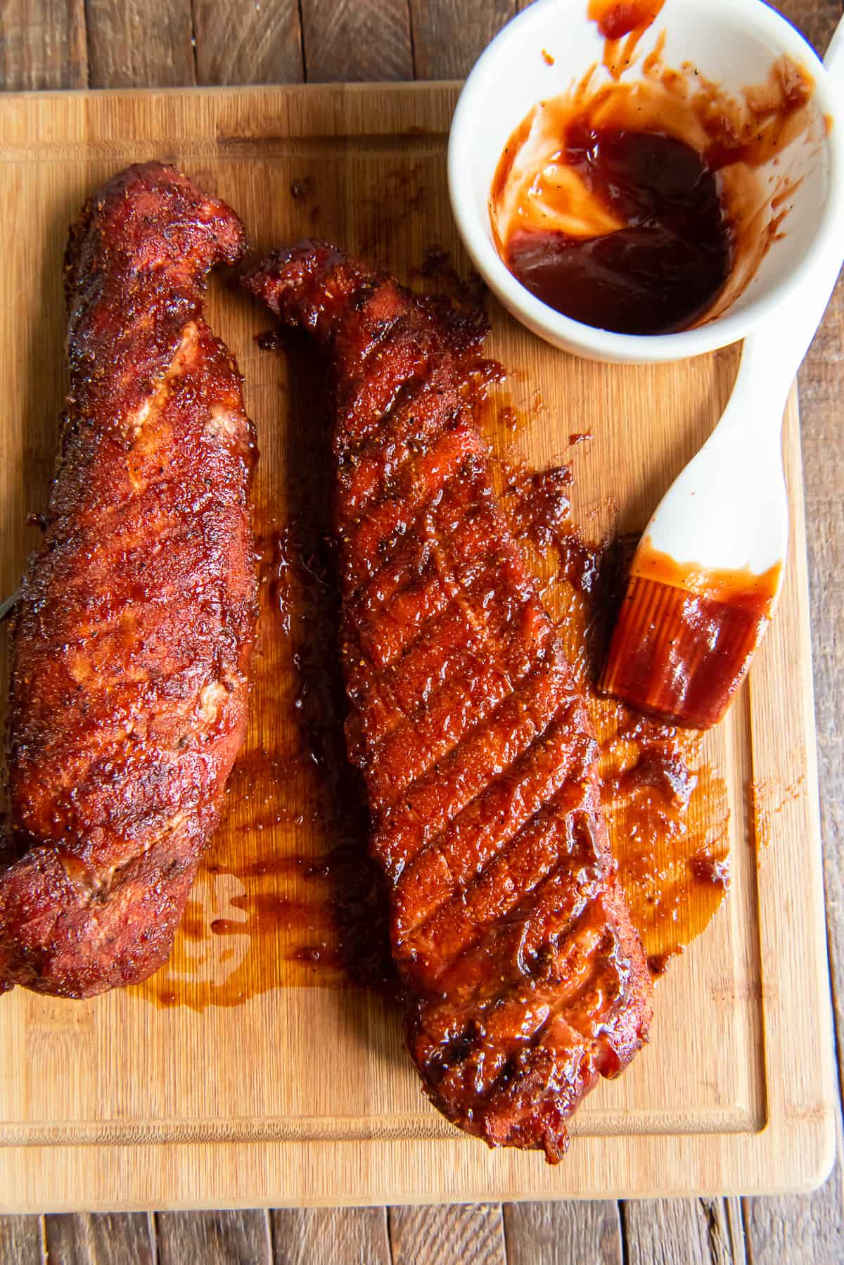Two smoked pork tenderloins on a cutting board with a bowl of barbecue sauce.