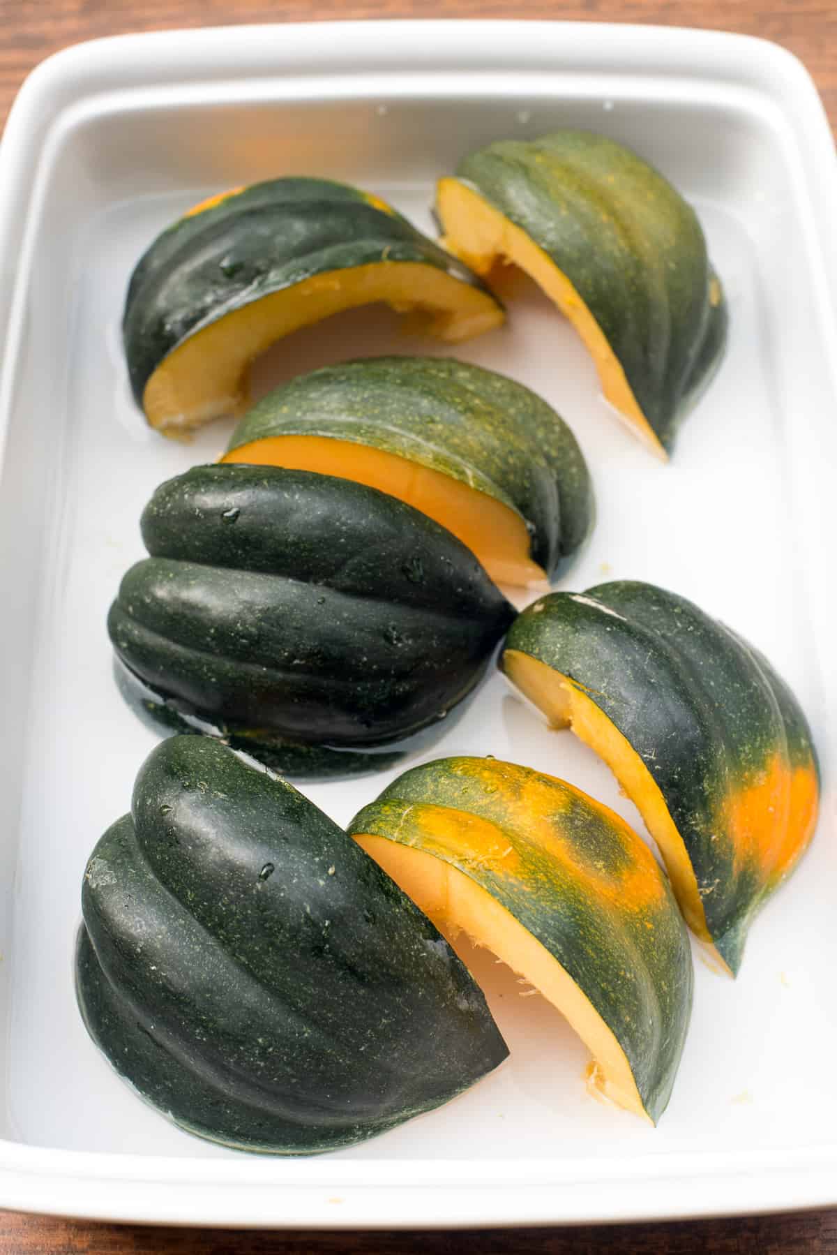 Quartered acorn squash placed cut side down in a white baking dish.