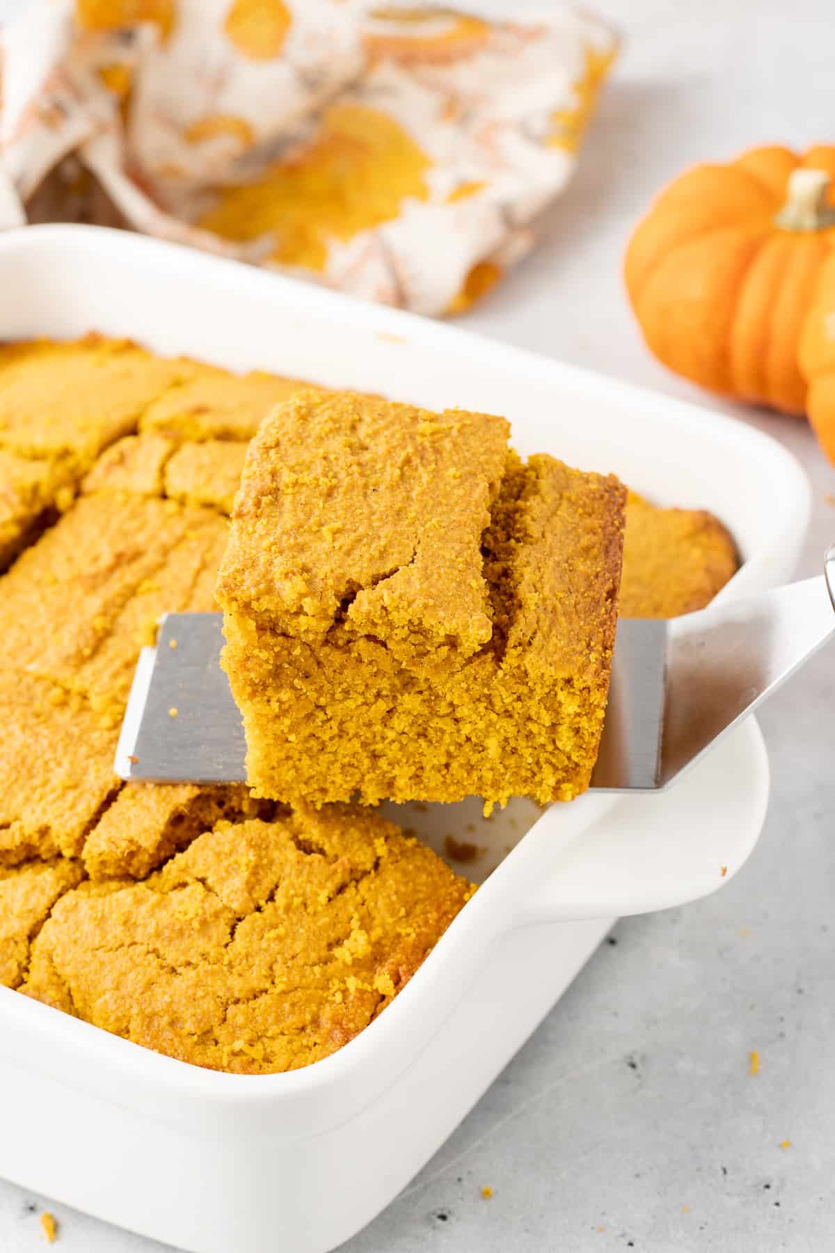 A spatula lifts a slice of pumpkin cornbread from a white baking dish.