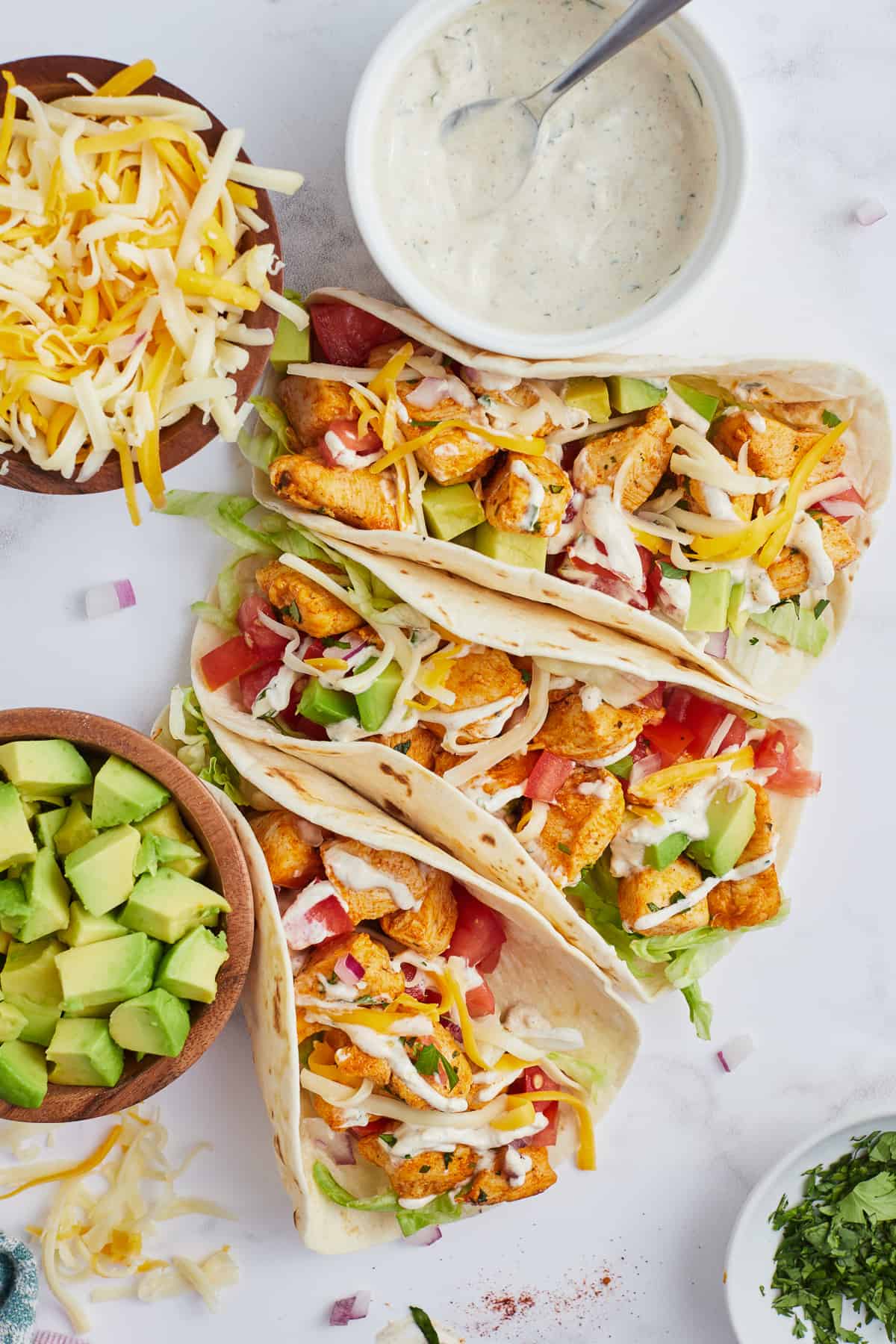 A top down shot of three chicken tacos next to a bowl of Ranch sauce, cheese, and avocado.