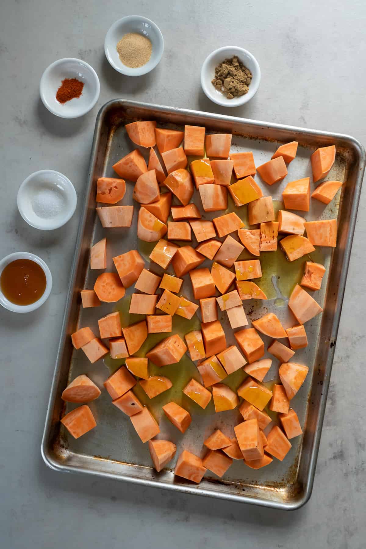 A top down shot of chunks of roasted sweet potatoes tossed with olive oil on a baking sheet with small bowls of seasoning around it.