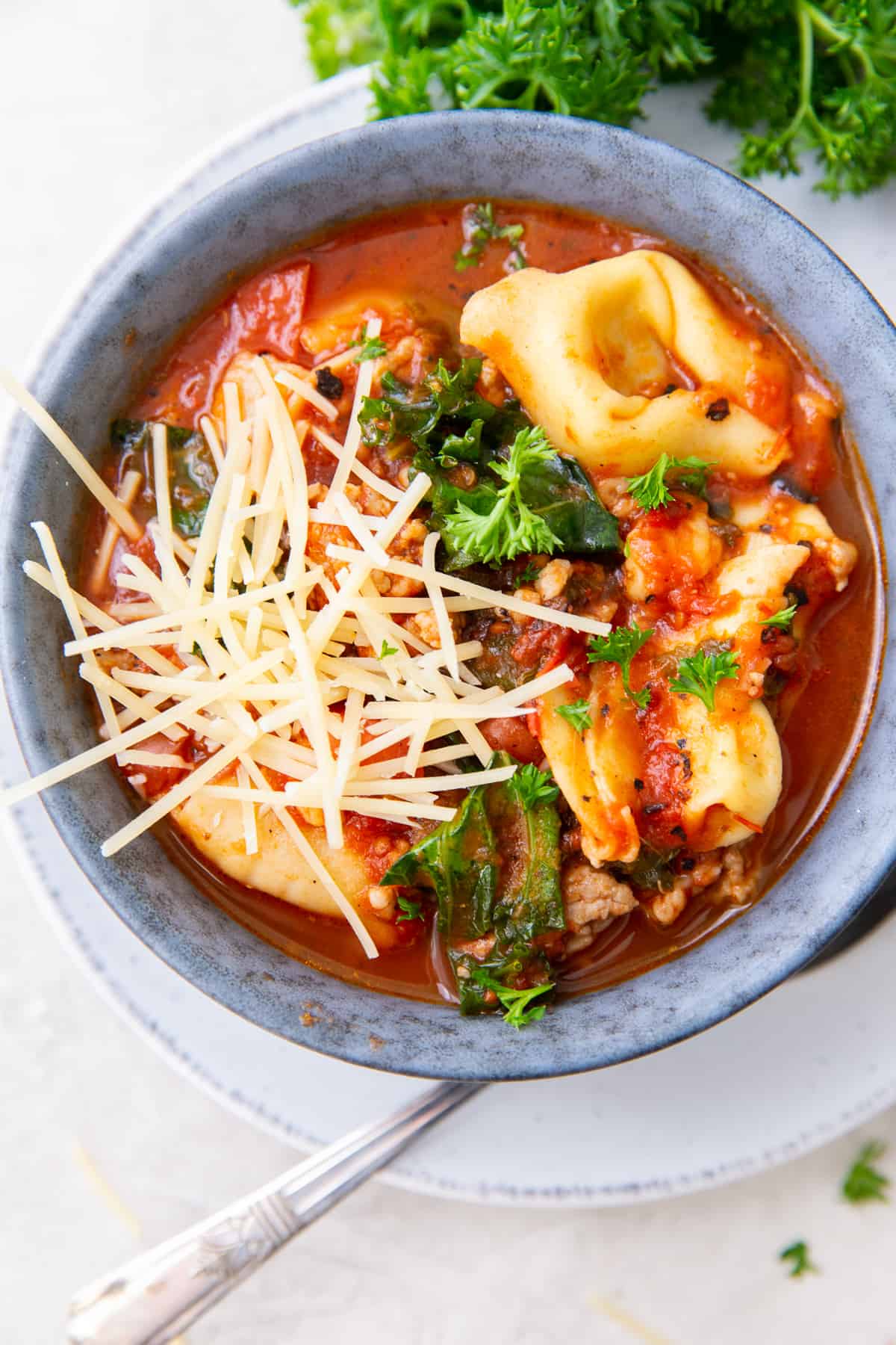 A top down shot of a bowl of soup with tortellini, sausage, kale and parmesan cheese.