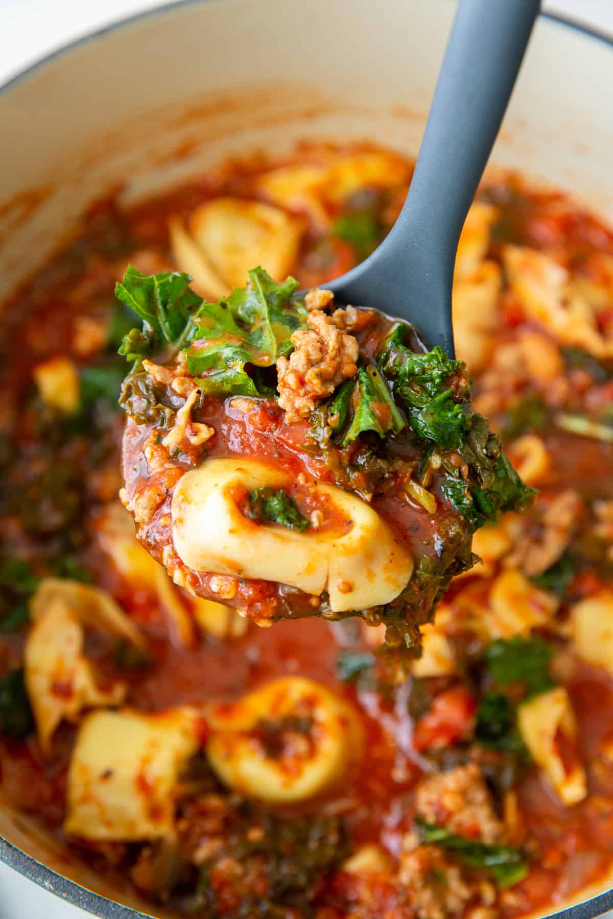 A ladle scooping up sausage tortellini soup with kale from a Dutch oven.