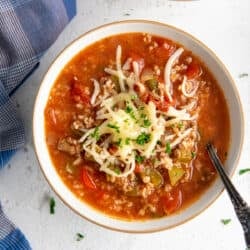 A top down shot of spoon resting in a bowl of soup with ground beef, rice, peppers, and topped with shredded cheese.