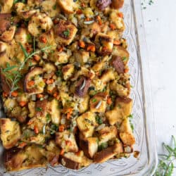 A top down shot of stuffing made with brioche in a glass baking dish surrounded by fresh herbs.