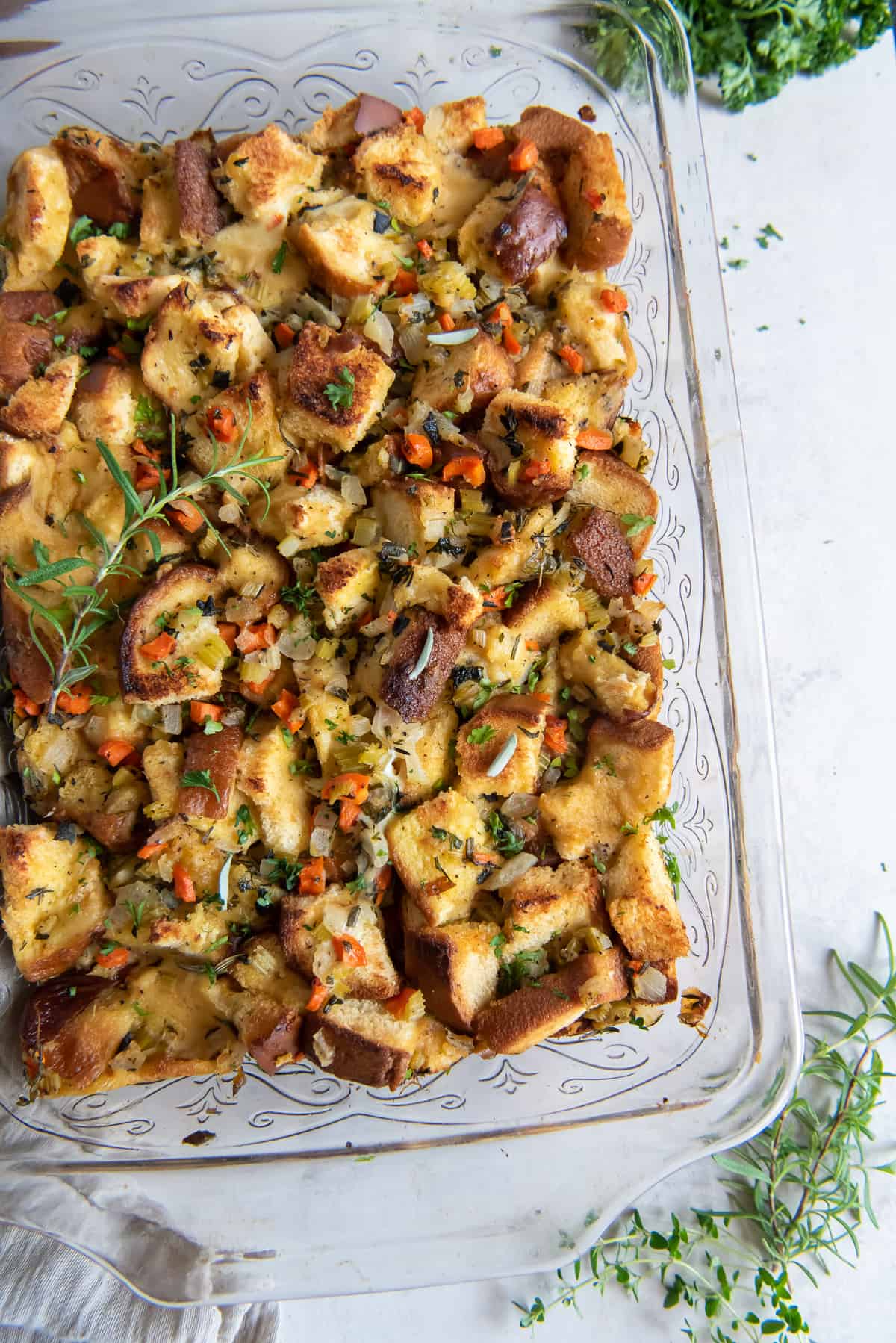 A top down shot of stuffing made with brioche in a glass baking dish surrounded by fresh herbs.