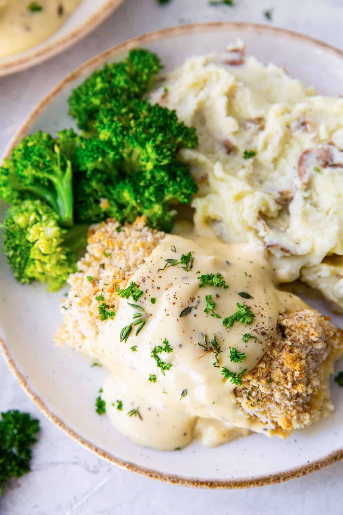 A breaded chicken roll topped with creamy sauce on a white plate with broccoli and mashed potatoes.