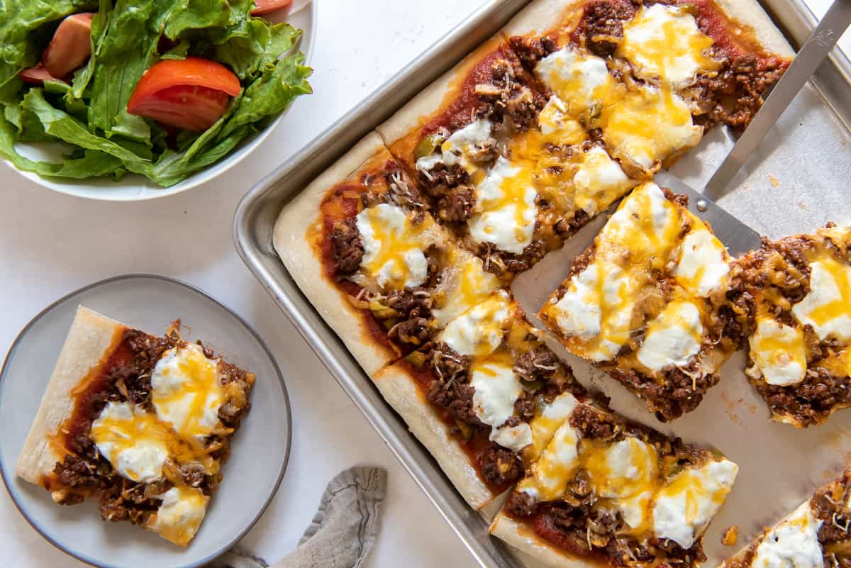 A top down shot of a cheeseburger sheet pan pizza next to a plate with one slice and green salad.