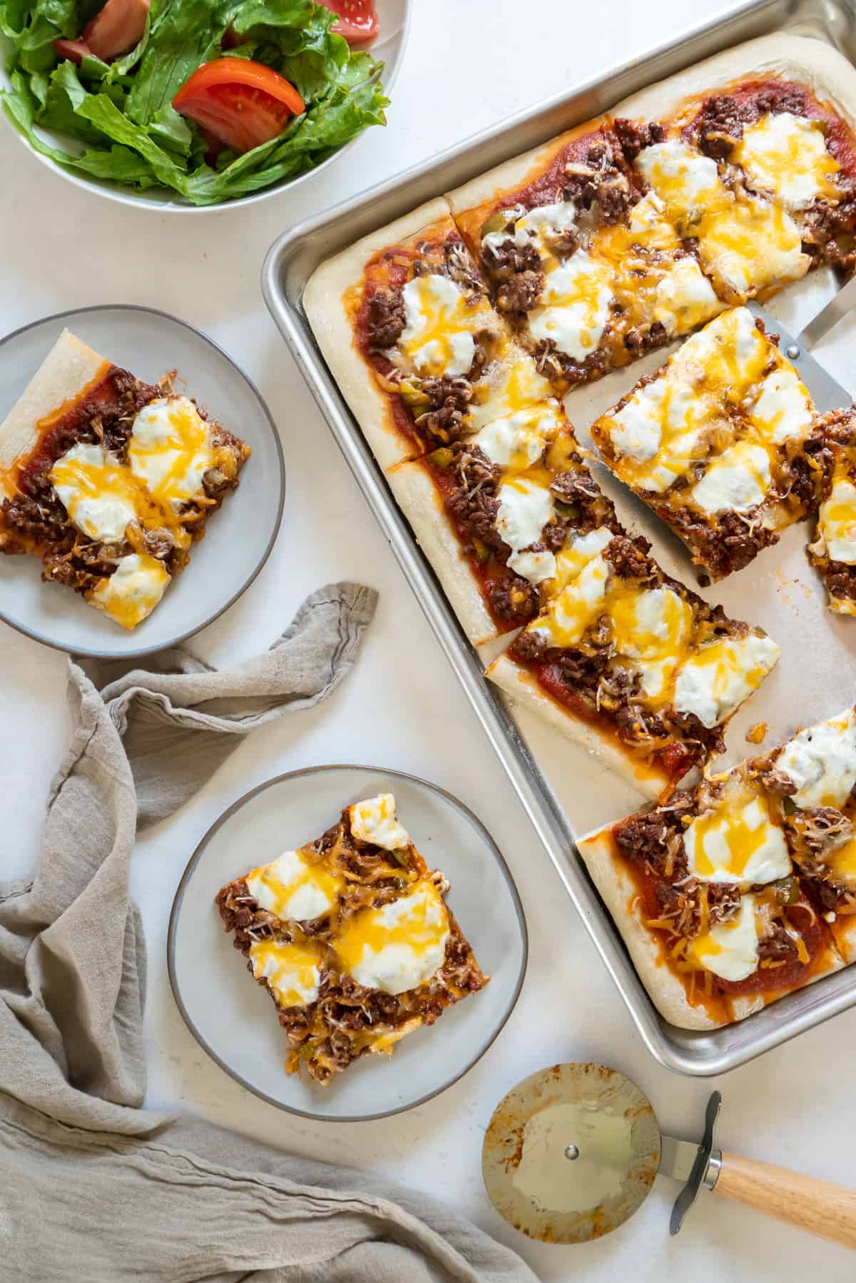 Two plates with slices of pizza and a green salad next to a cheeseburger pizza on a baking sheet.