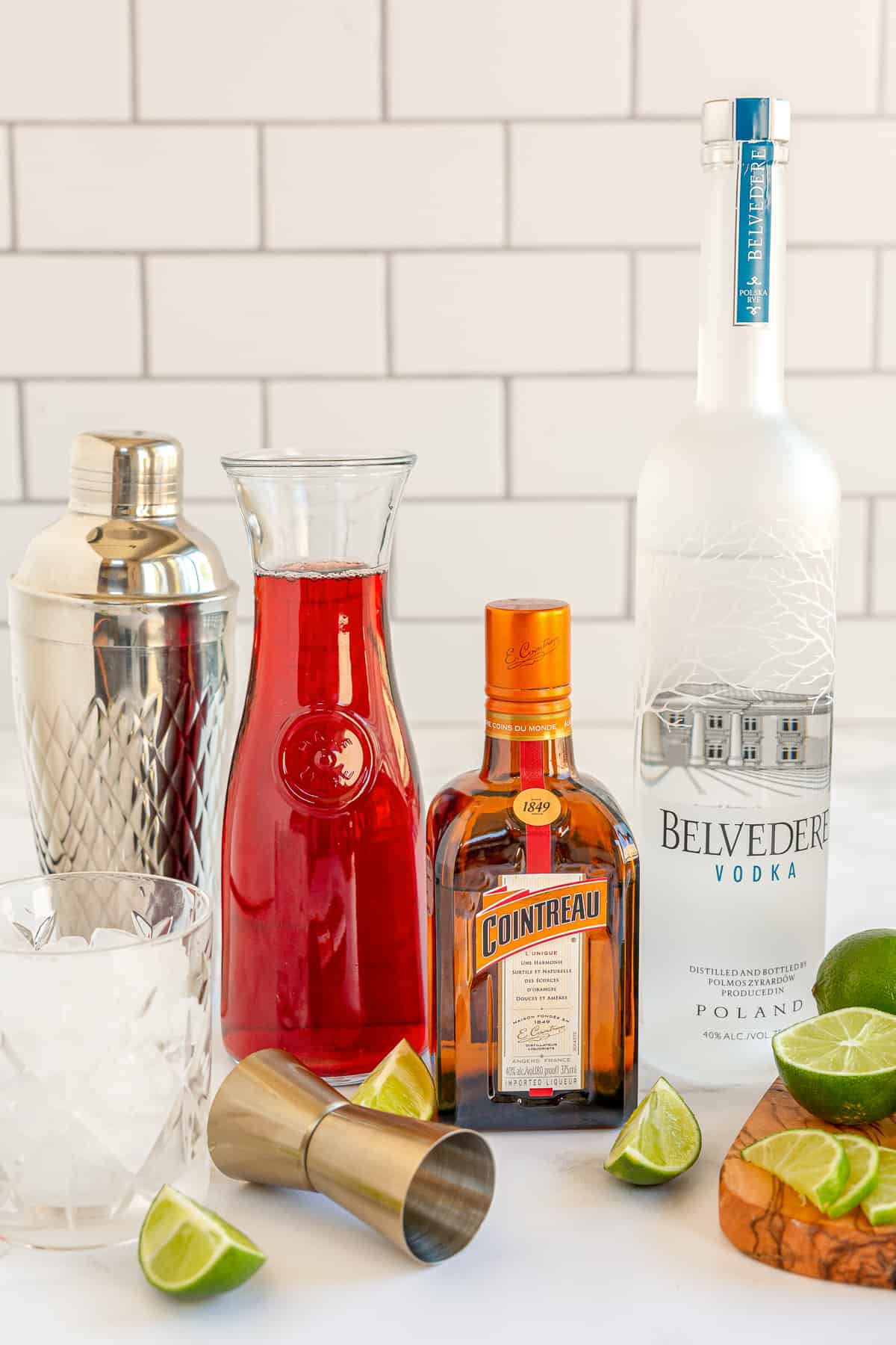 A bottle of vodka, Countreau, and a carafe of cranberry juice surrounded by a cocktail shaker and lime wedges.