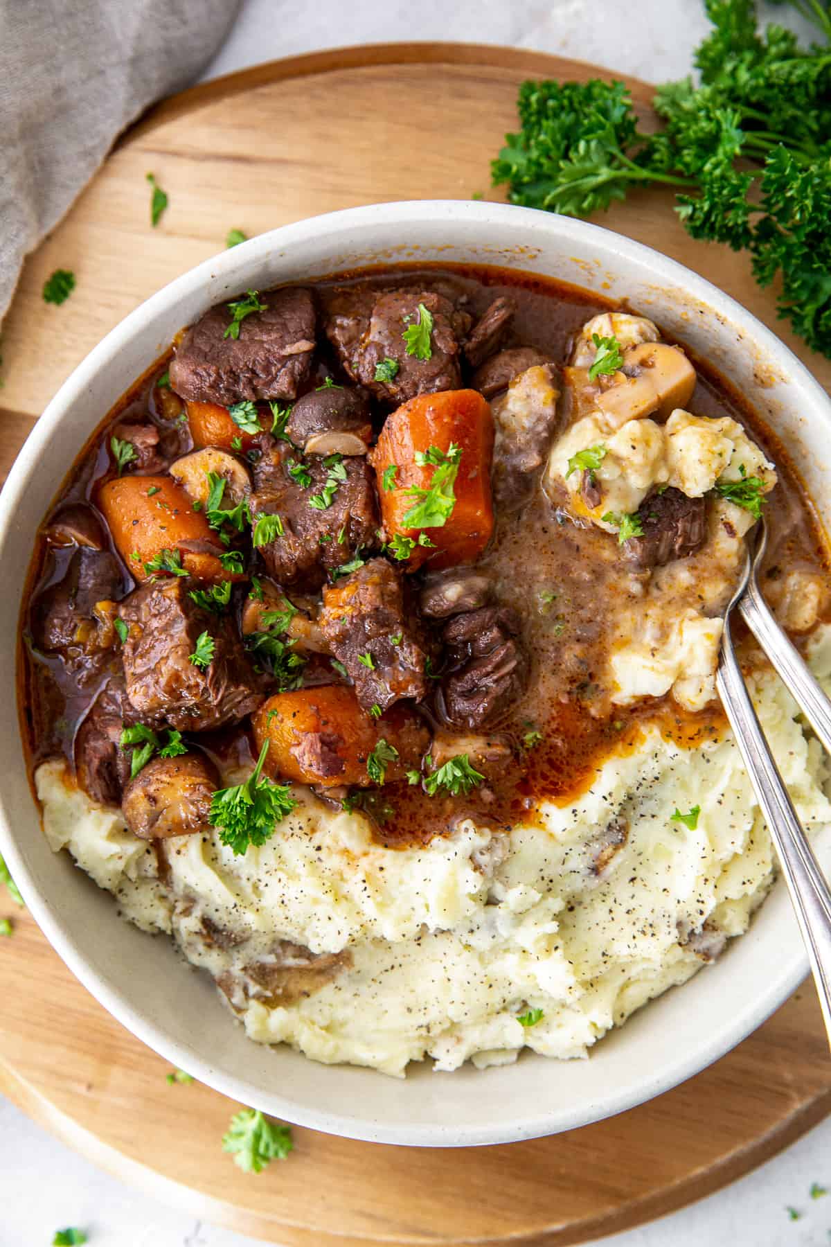 A top down view of cubes of beef, carrots, and mushrooms in gravy over mashed potatoes in a white bowl on a board.