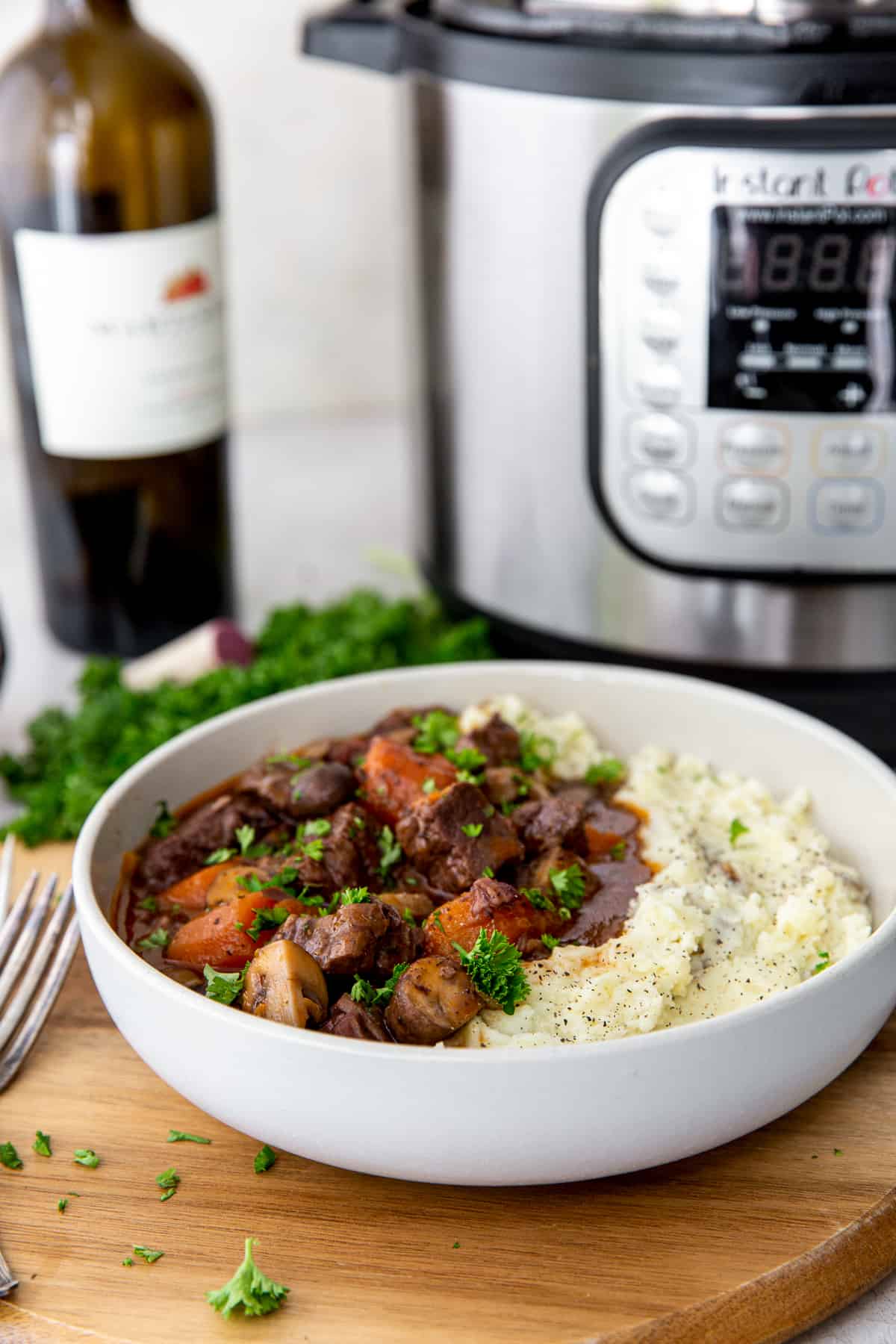 A white bowl with beef burgundy and mashed potatoes in front of an Instant Pot. and a bottle of wine.