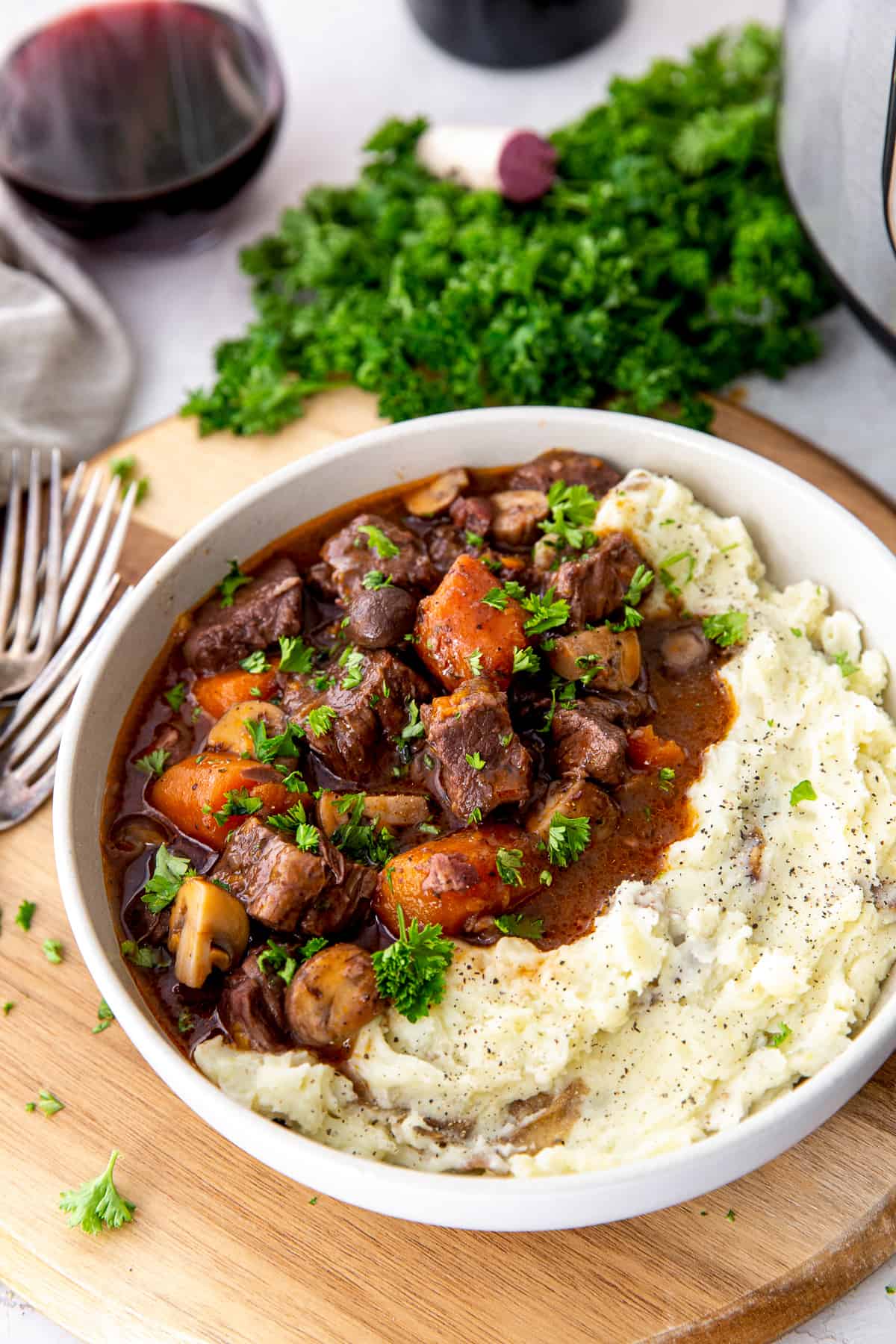 Beef burgundy with carrots and mushrooms in a white bowl with mashed potatoes in front of a glass of wine.