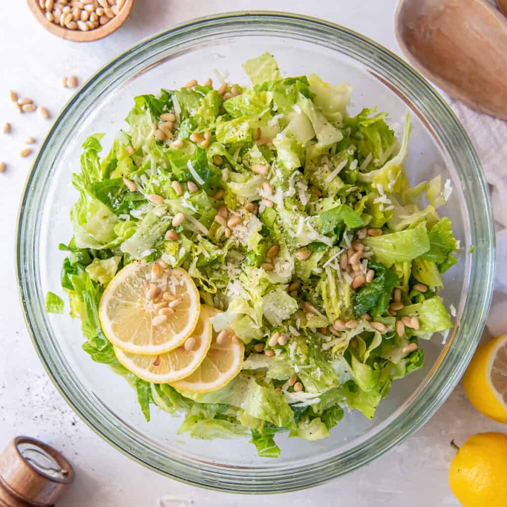 A top down shot of a salad of chopped romaine with Parmesan cheese and pine nuts garnished with lemon slices.