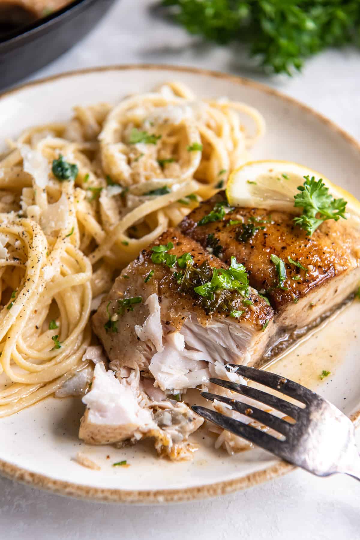 A fork breaking apart a tender and flaky mahi mahi fillet on a white plate with pasta.