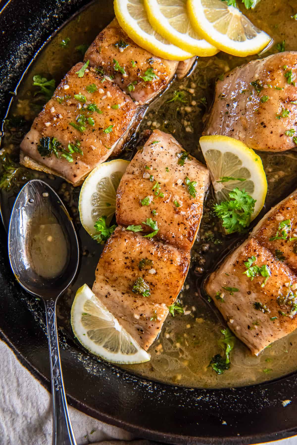 A close up top down shot of mahi mahi with sauce and lemon in a cast iron skillet with a metal spoon.