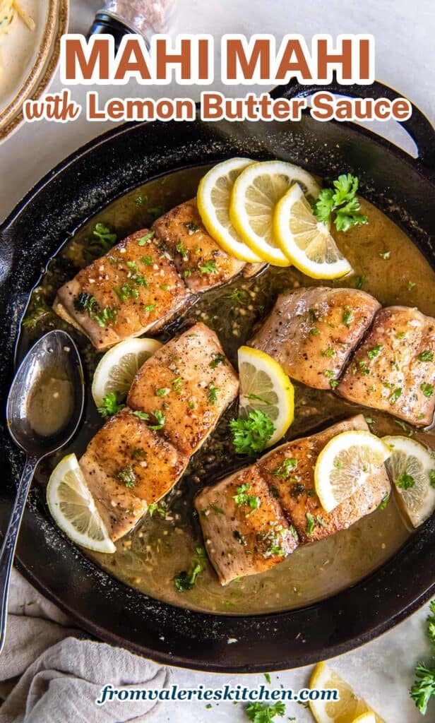 A top down shot of mahi mahi with sauce and lemon in a cast iron skillet with a metal spoon with text.