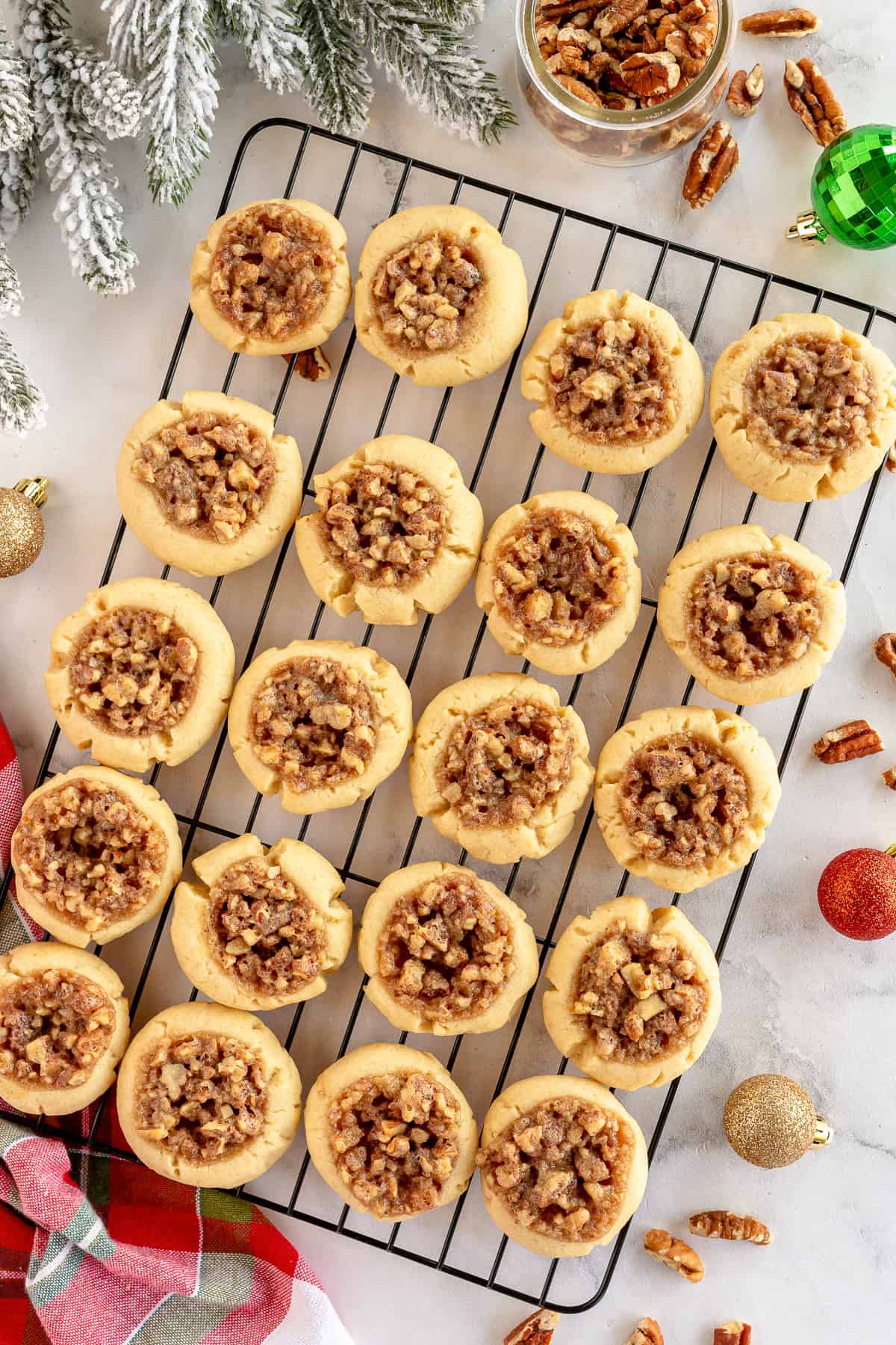 Thumbprint cookies with a pecan mixture on a wire cooling rack with Christmas ornaments and pine tree branches around the edges.