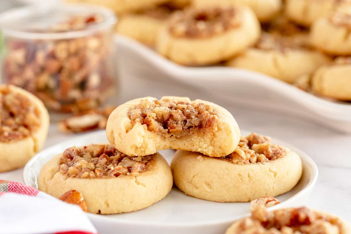 A Pecan Pie Thumbprint Cookie with a bite missing stacked on two cookies on a white plate.