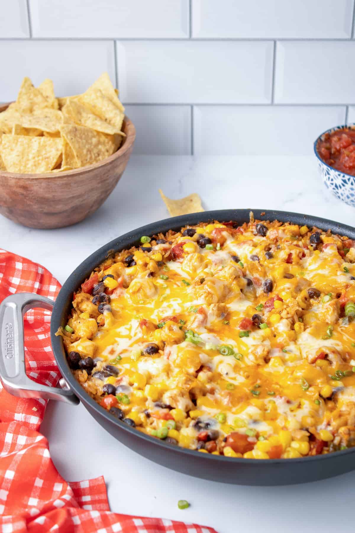 A skillet full of tex mex chicken and rice with melted cheese on a kitchen counter with tortilla chips and salsa in the background.