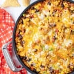 A top down shot of a skillet filled with a chicken and rice mixture with melted cheese next to a red and white checkered cloth.