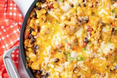 A top down shot of a skillet filled with a chicken and rice mixture with melted cheese next to a red and white checkered cloth.