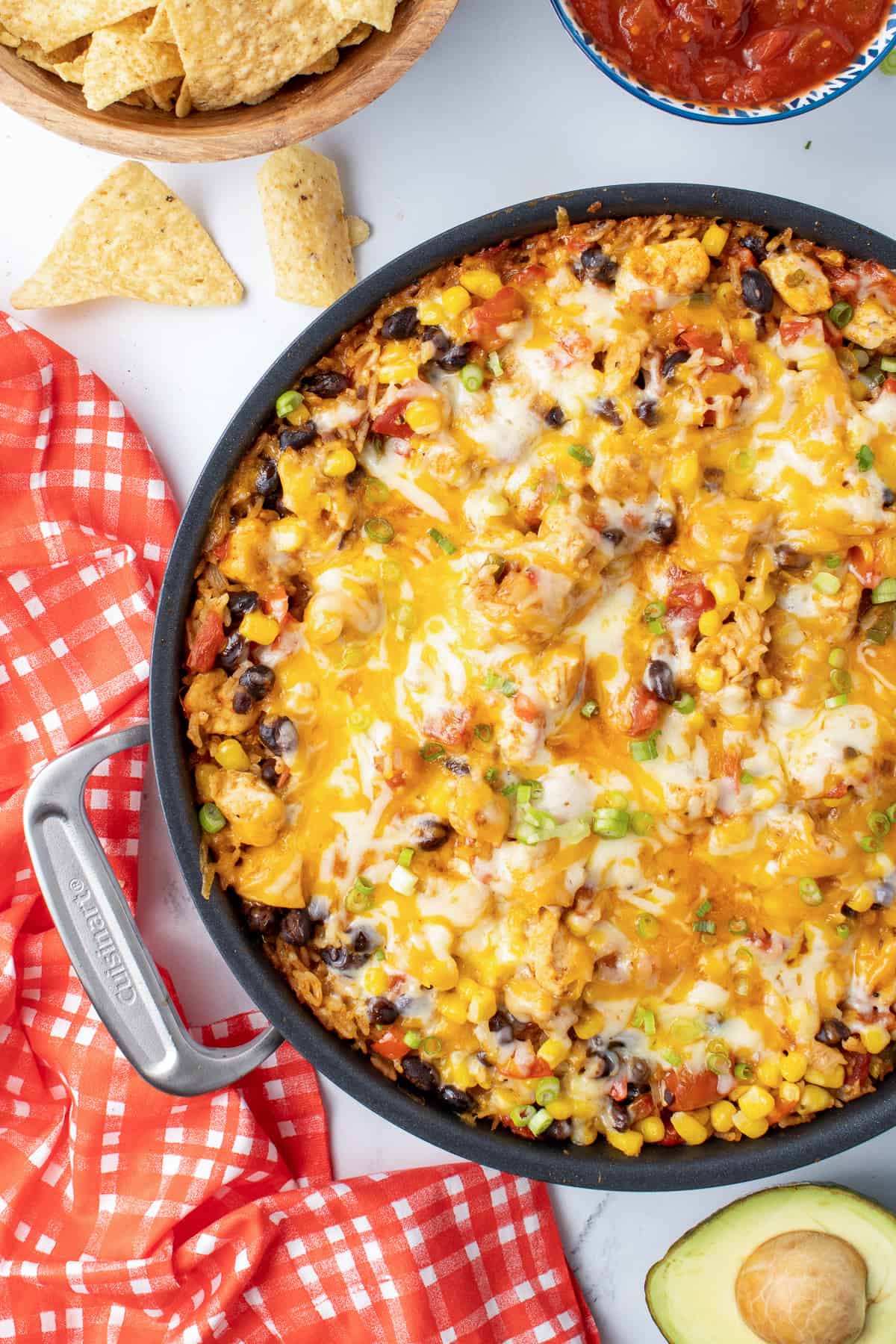 A top down shot of a skillet filled with a chicken and rice mixture with melted cheese next to a red and white checkered cloth.