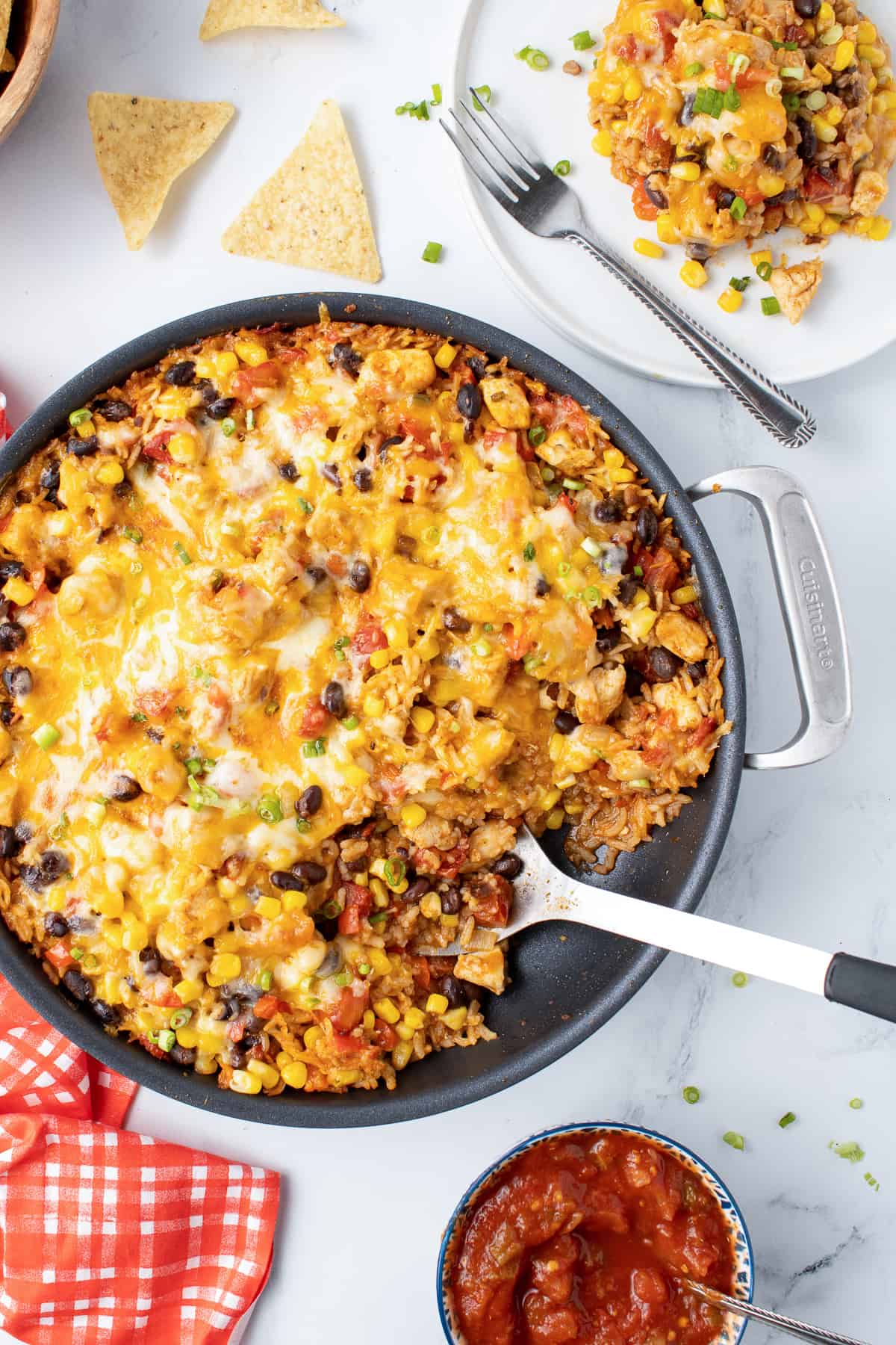 A spoon resting in a skillet filled with cheesy tex mex chicken and rice next to a plate with a fork resting on it.