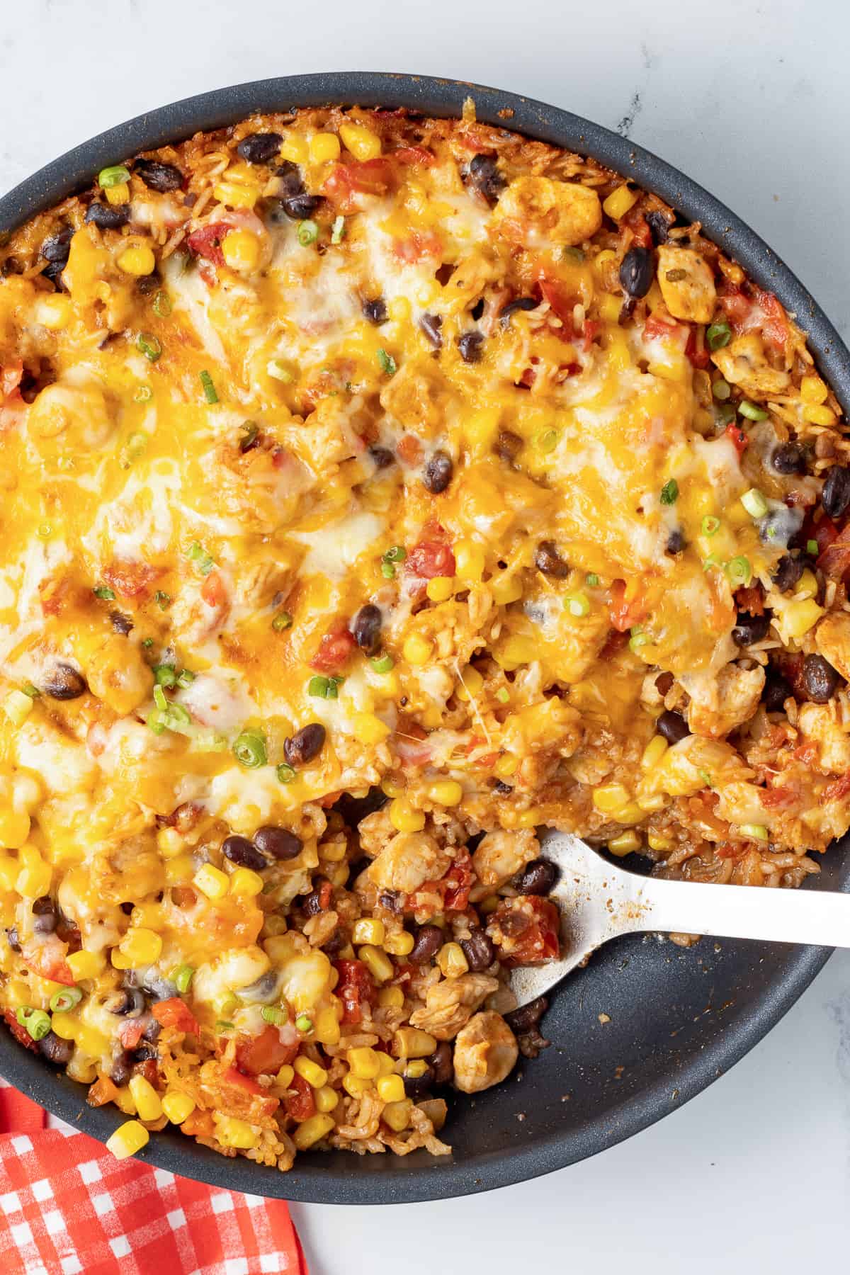 A spoon resting in a skillet filled with cheesy tex mex chicken and rice next to a red and white checkered cloth.