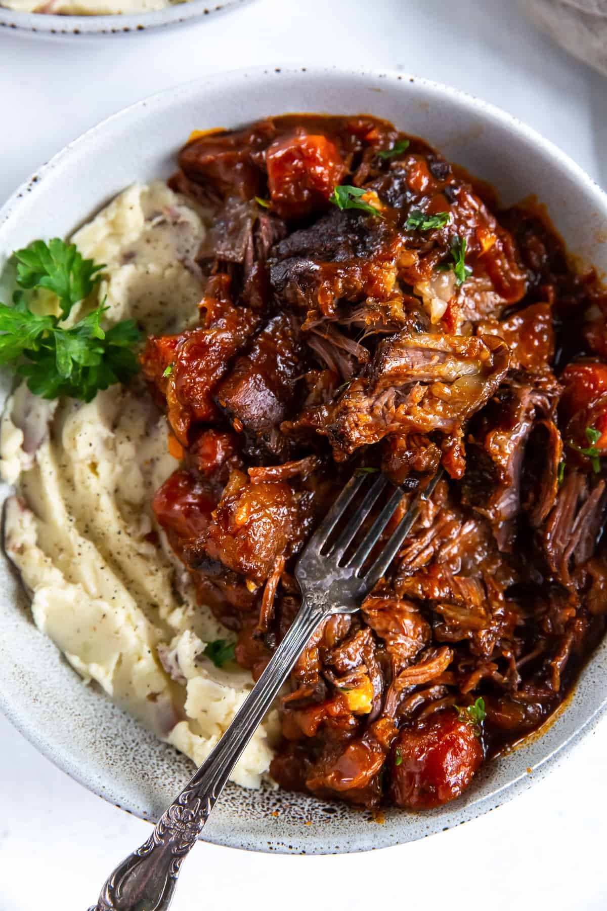 A top down shot of a fork resting in a bowl of short ribs in a saucy bbq mixture and mashed potatoes.