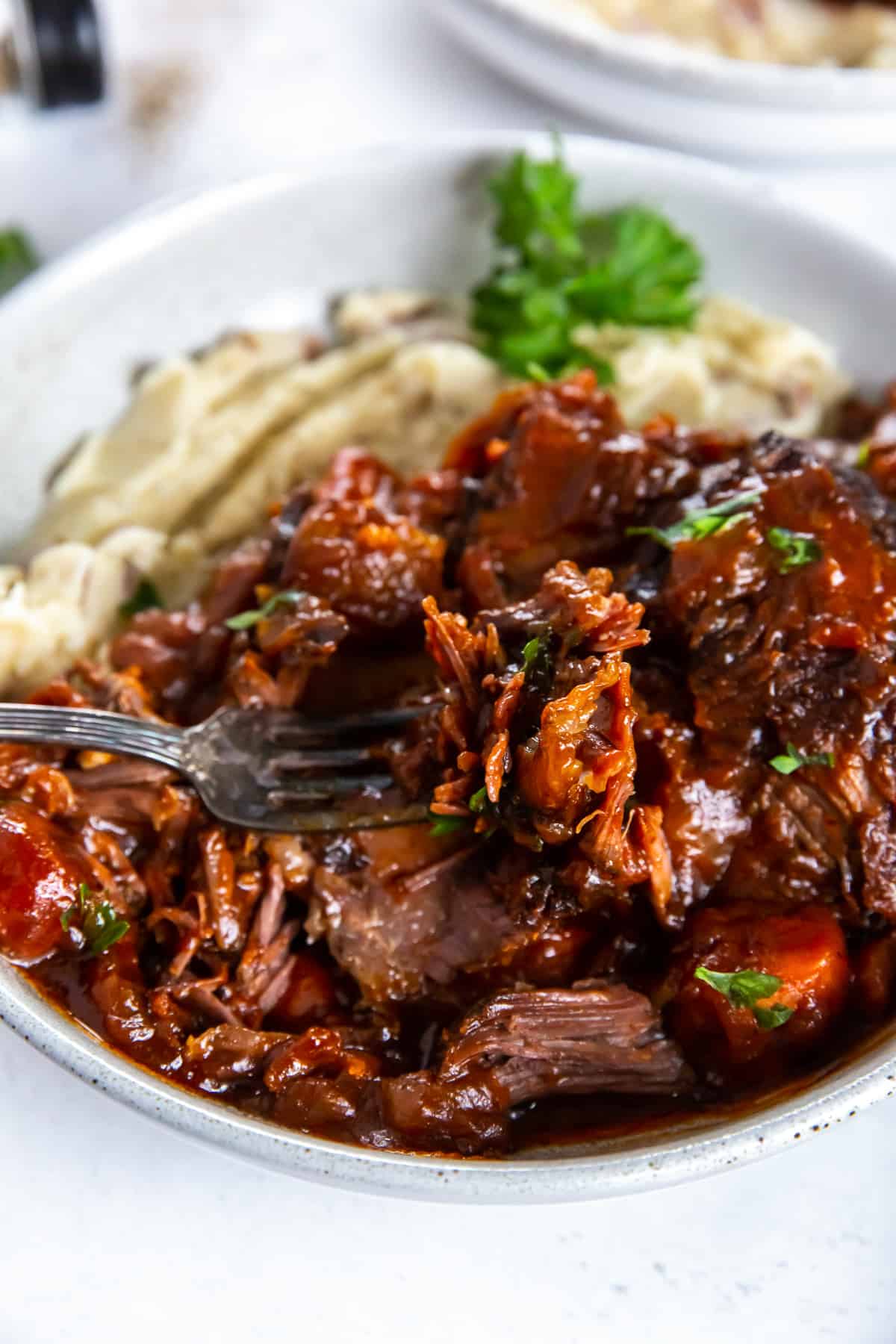 A fork filled with tender braised short rib meat hovering over a white bowl.