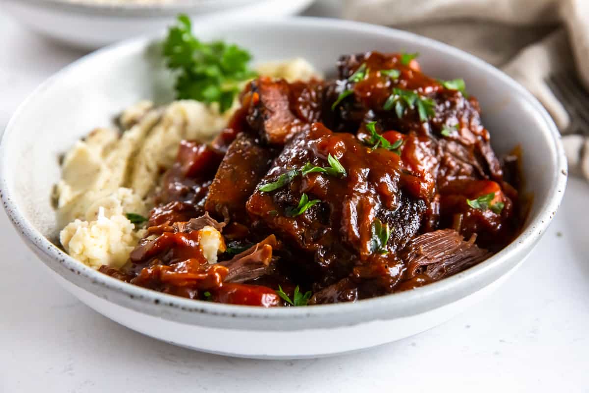 A side view of a white bowl filled with short ribs in a saucy BBQ mixture and mashed potatoes.