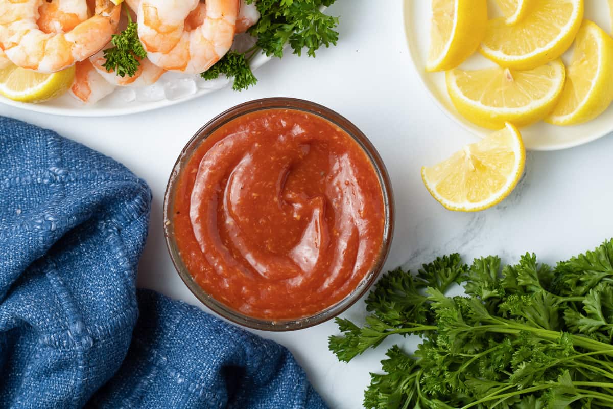 A top down shot of cocktail sauce in a bowl next to a blue cloth, a platter of shrimp, and lemon wedges.