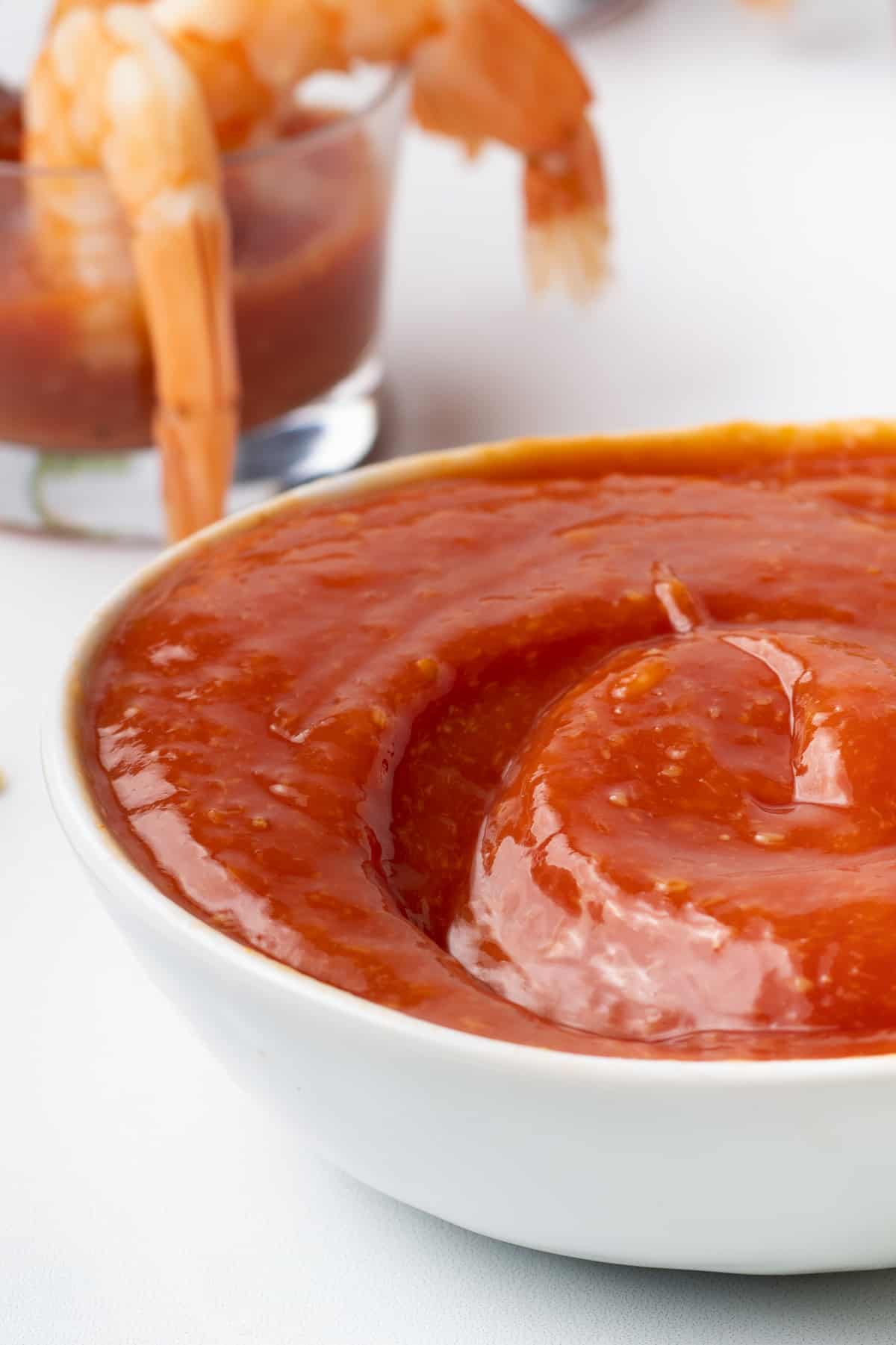 A close up side view of cocktail sauce in a white bowl with shrimp in the background.