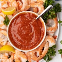 A top down shot of cocktail sauce in a small bowl with a spoon on a platter full of shrimp.