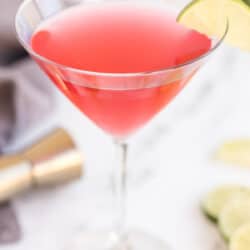 A cosmopolitan cocktail in a martini glass with a slice of lime on the rim on a kitchen counter.