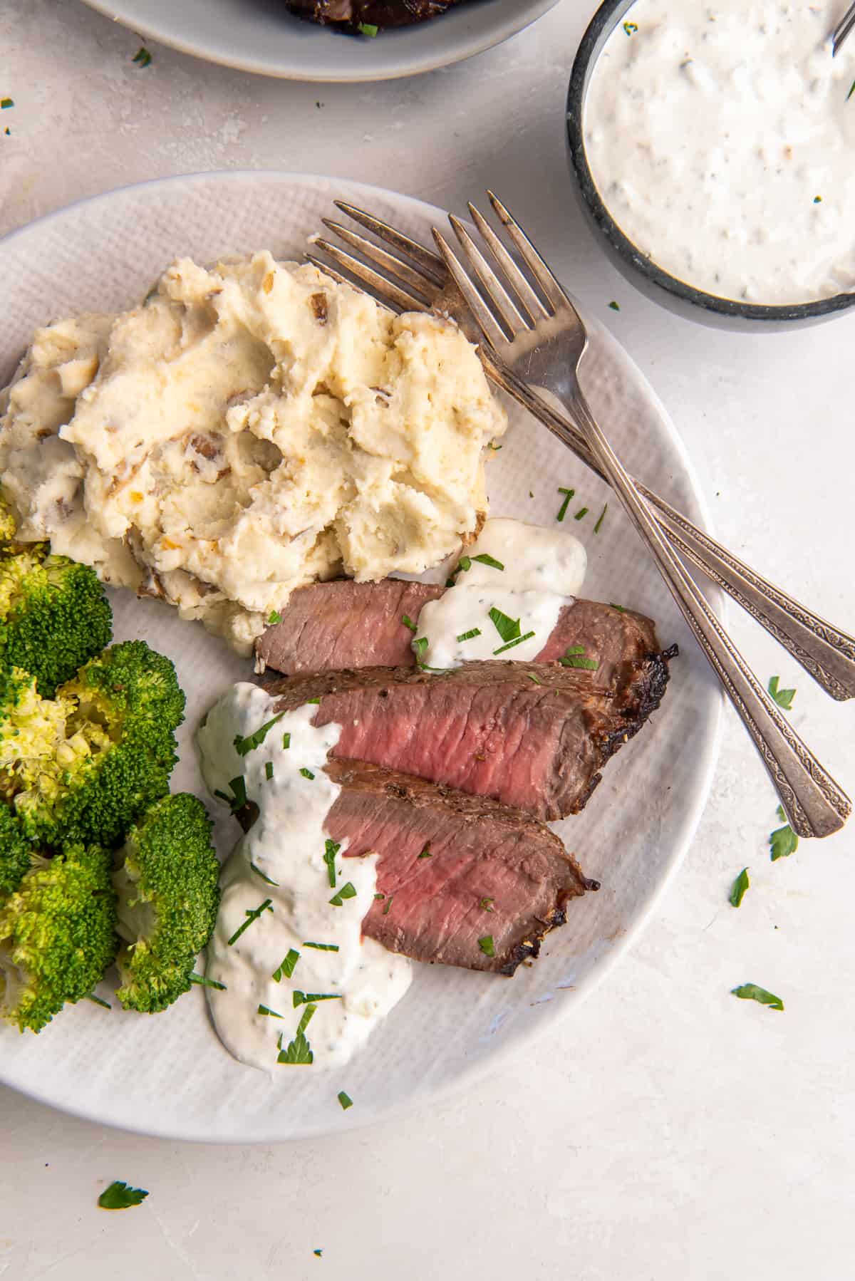 Slices of London Boil topped with a creamy sauce on a white plate with broccoli and mashed potatoes.