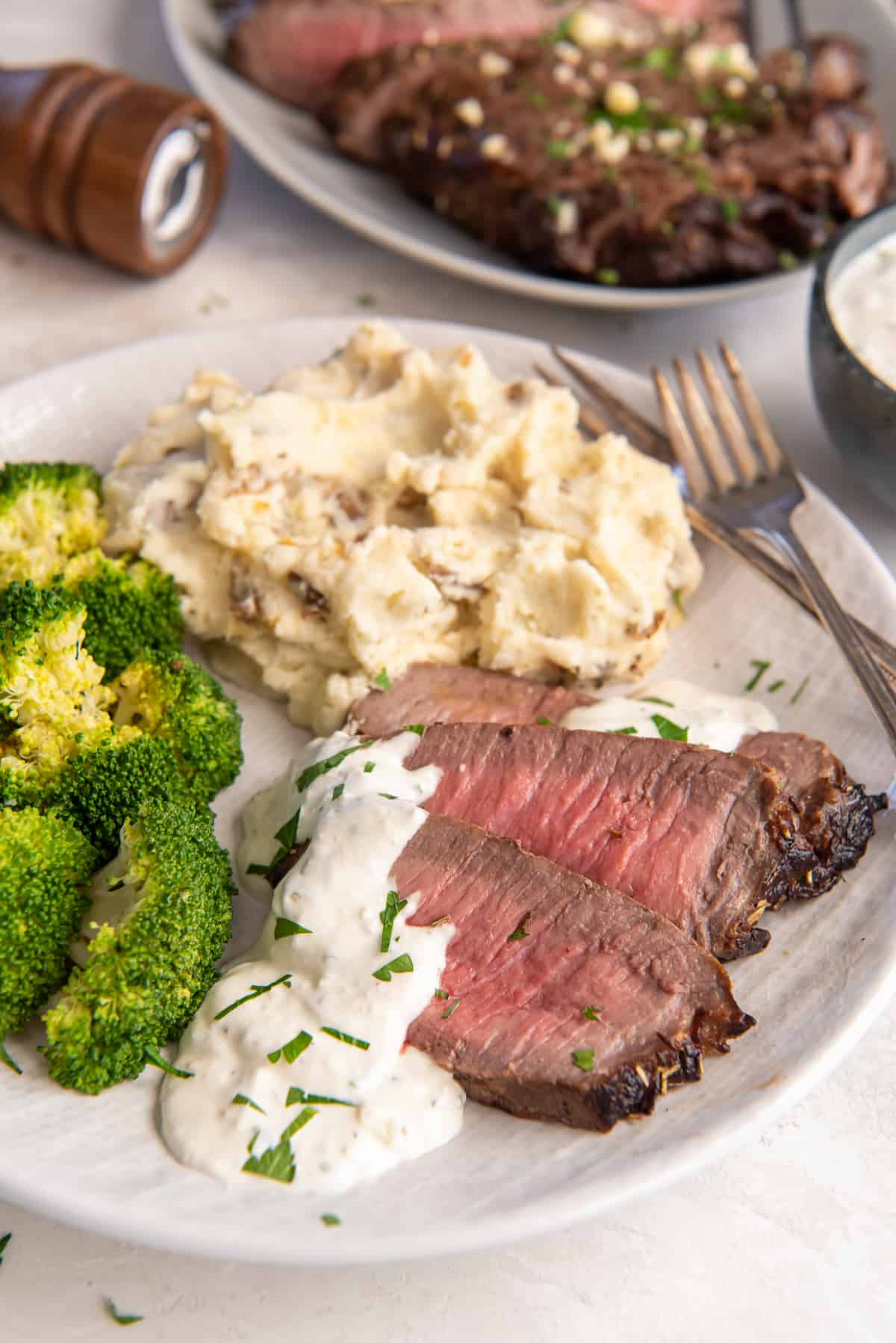 Slices of London Boil topped with a creamy sauce on a white plate with broccoli and mashed potatoes.